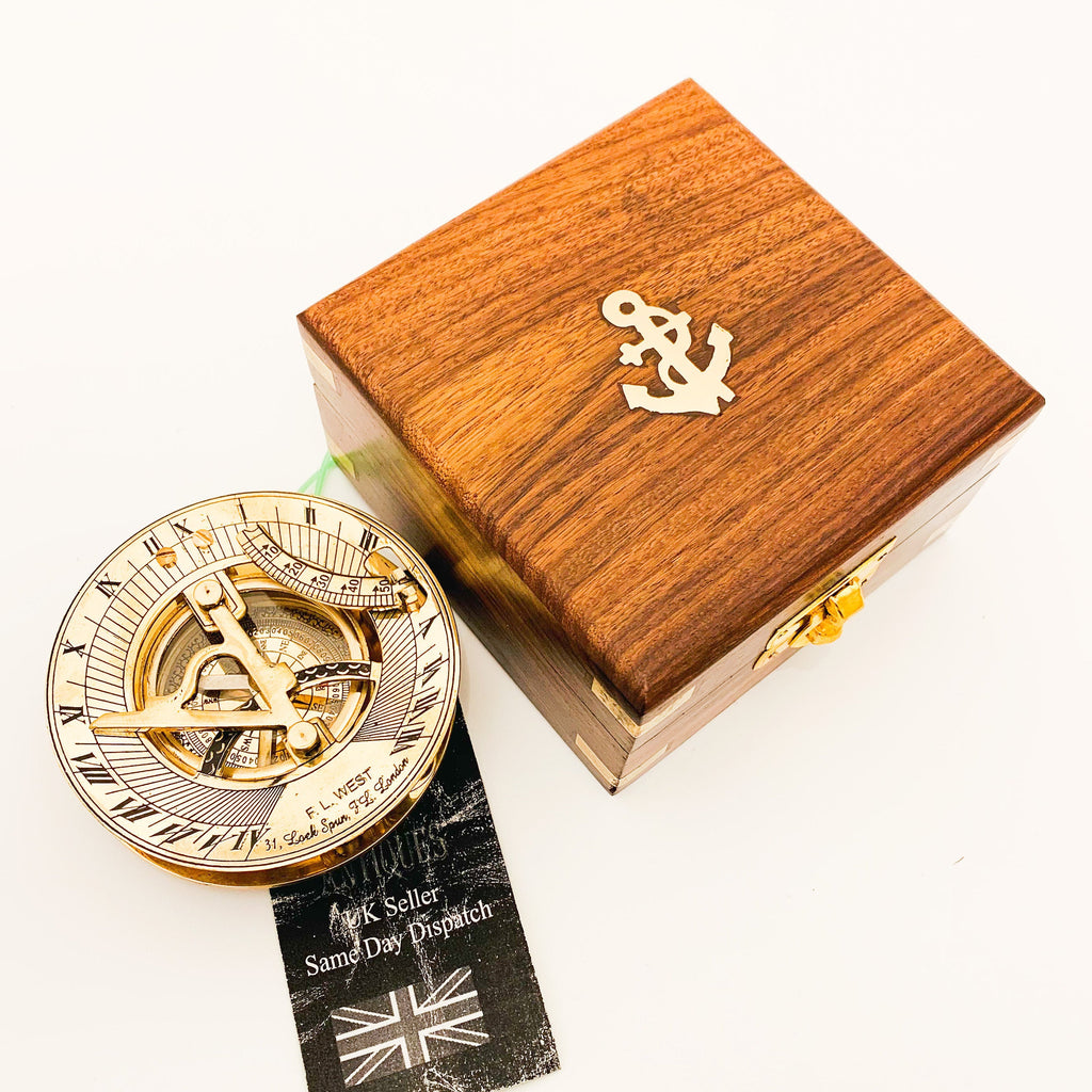May include: A brass sundial compass with a wooden box. The compass has a compass rose, a sundial, and a protractor. The box is made of dark wood and has an anchor design on the top.