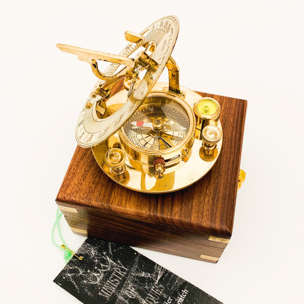 May include: A brass compass and sundial set in a wooden box. The compass has a white face with black markings and a red needle. The sundial has a gold finish and is decorated with intricate details.