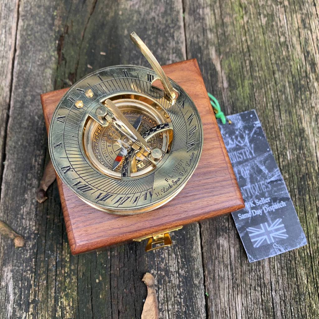 Puede incluir: Un reloj de sol de latón antiguo con números romanos y una brújula, en una caja de madera. El reloj de sol tiene un gnomon y está marcado con el texto "F.L. WEST 31. Lach Spur, Bus London". Una etiqueta con "MINISTRY OF ANTIQUES" está adjunta.