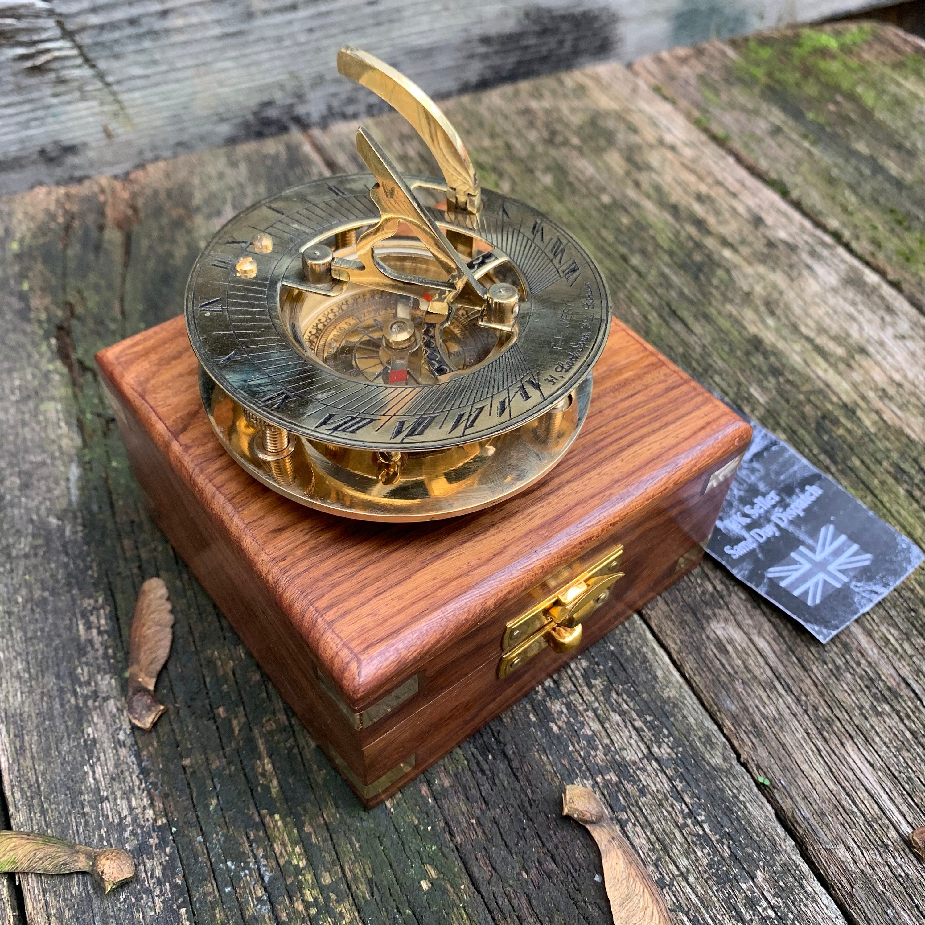 Puede incluir: Un reloj de sol de latón con brújula, colocado sobre una caja de madera. El reloj de sol tiene números romanos y un gnomon con bisagras. La caja es de madera oscura con herrajes de latón. Una etiqueta con el texto "UK Seller Same Day Dispatch" es visible.