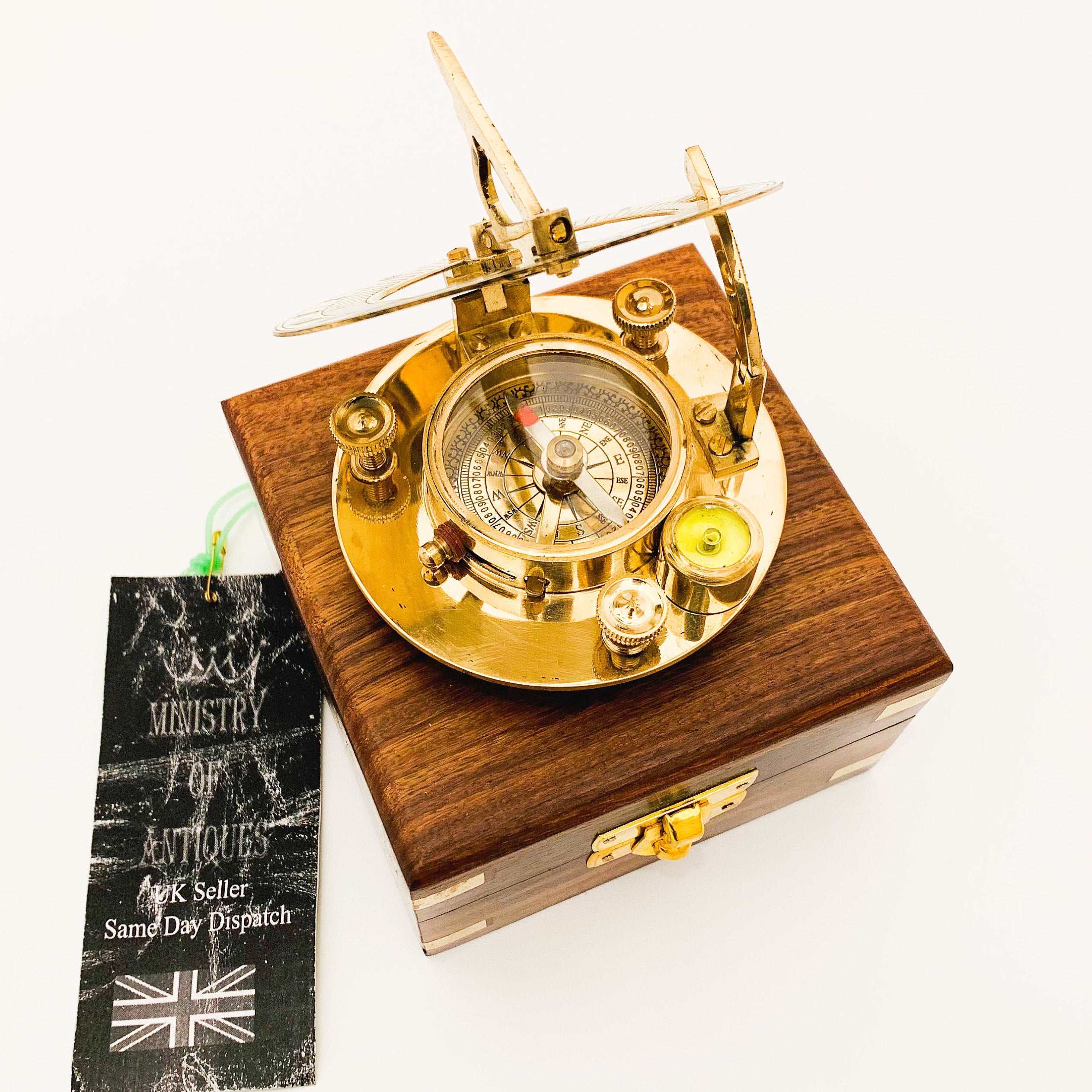 May include: A brass compass and sundial in a wooden box. The compass has a gold face with black markings and a red needle. The sundial has a brass arm and a white face with black markings. The box is made of dark wood with a brass latch.