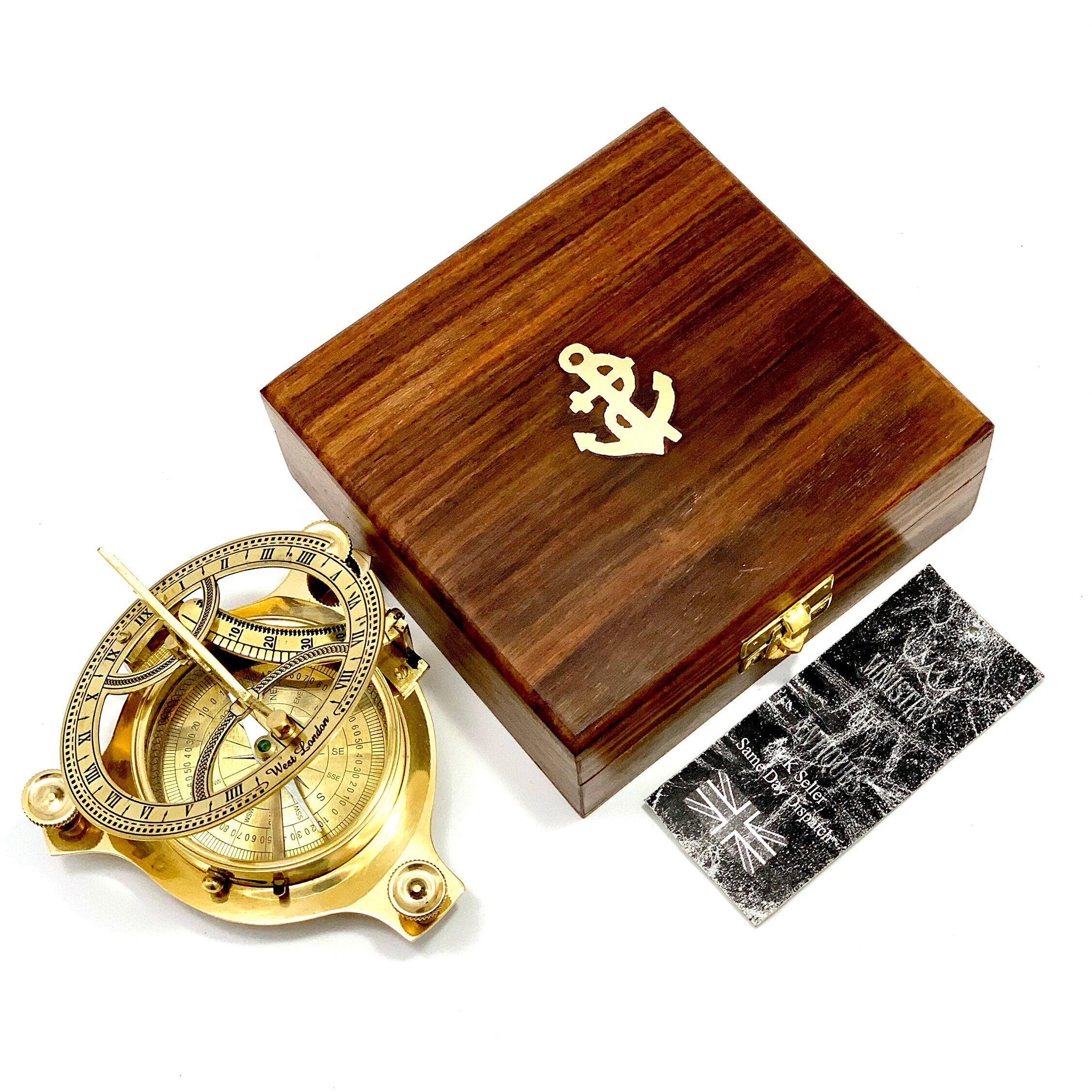 May include: A brass sundial compass with a wooden box. The compass features a compass rose, hour markings, and a gnomon. The box is made of dark wood and has an anchor design on the top.