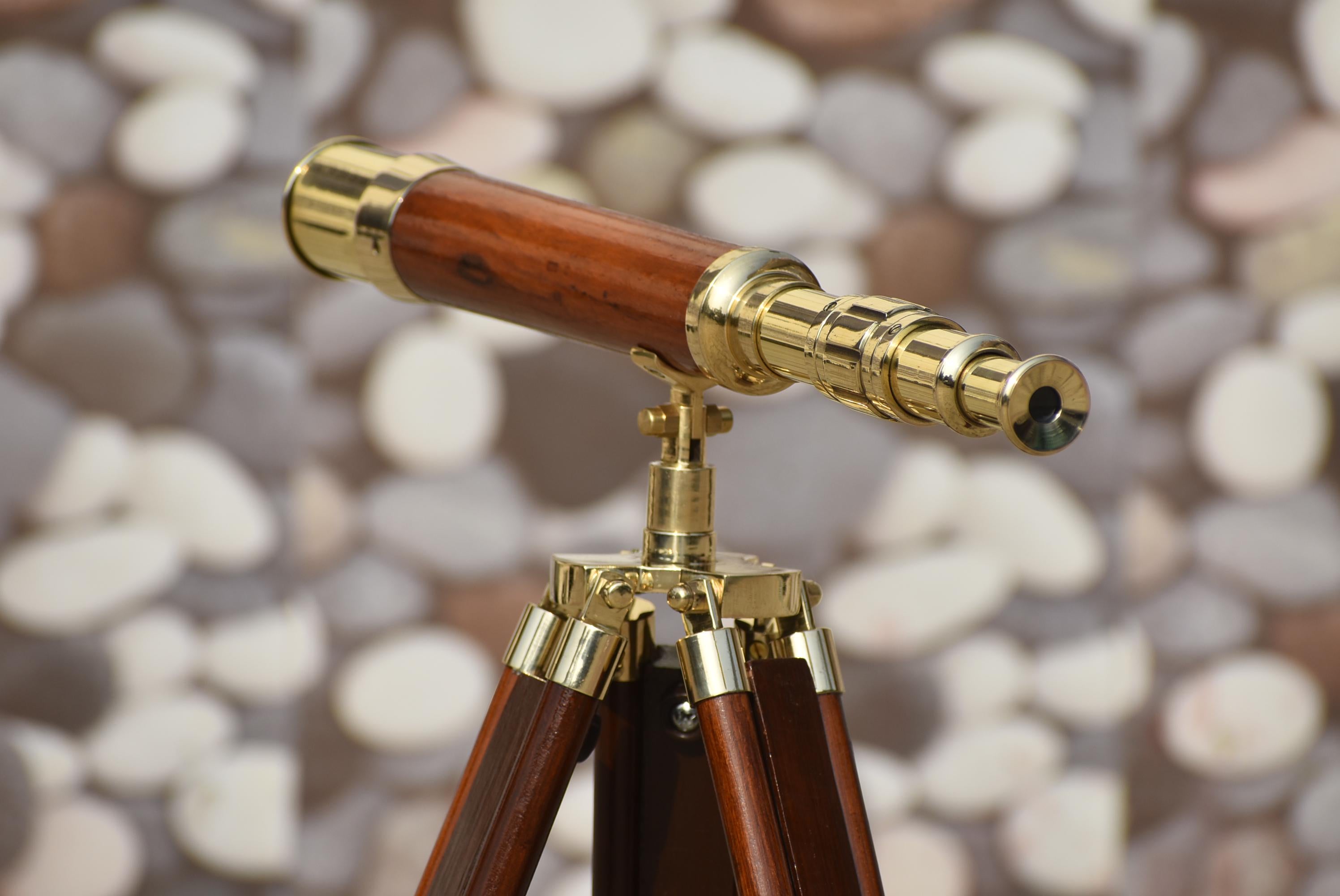 May include: A brass and wood telescope, reminiscent of a vintage design, is mounted on a wooden tripod. The telescope features a dark brown wooden barrel and brass detailing. The tripod legs are also dark brown wood with brass accents. The background is a blurred stone pattern.