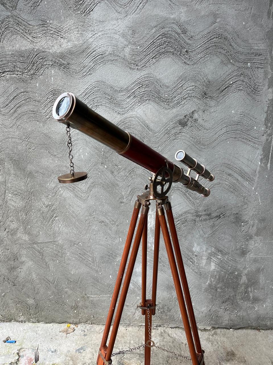 May include: A classic brass and wood telescope mounted on a wooden tripod. The telescope features a dark red and brass barrel and a small secondary scope. The tripod legs are a warm brown, set against a textured grey wall.