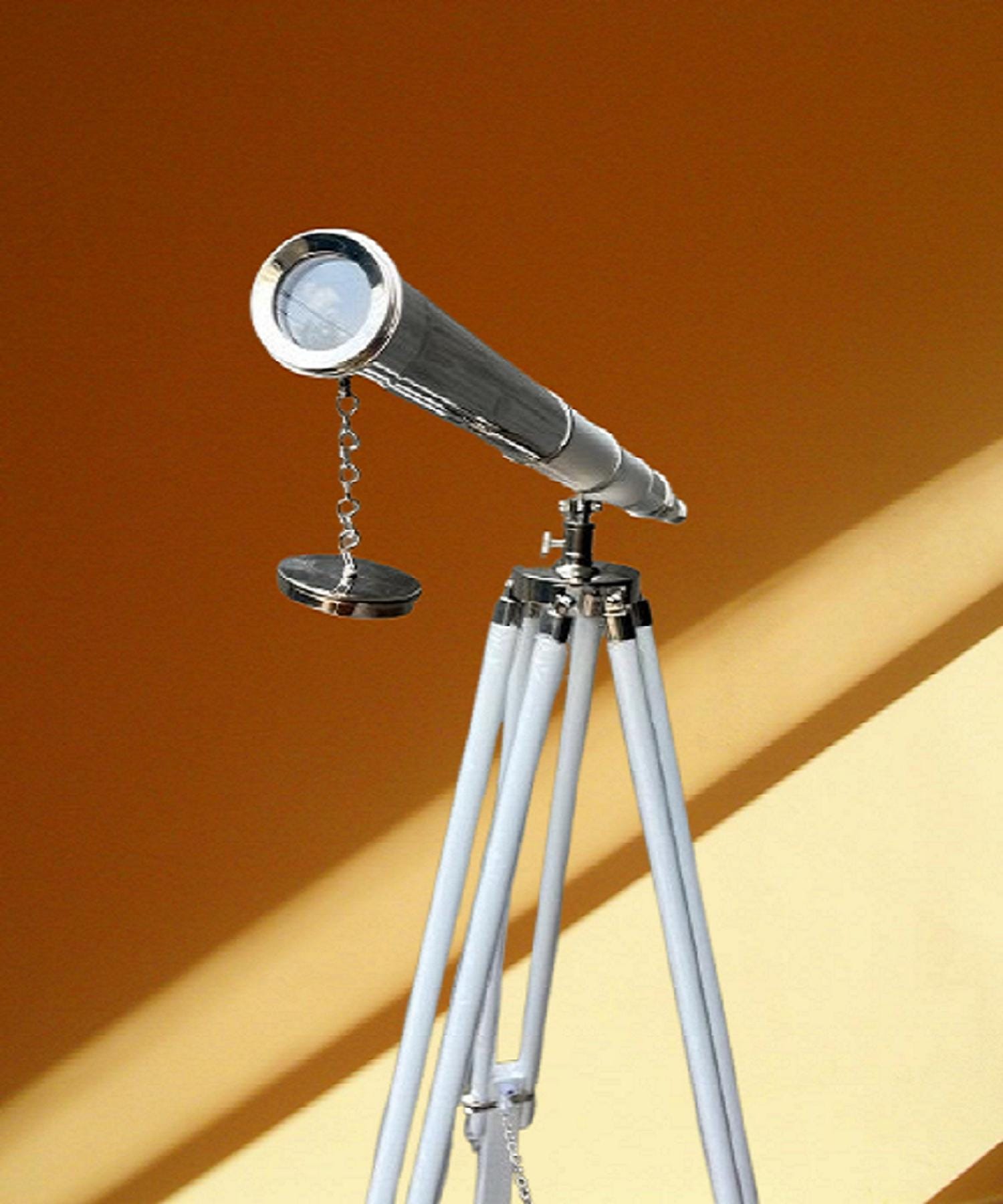 May include: A chrome telescope on a white and silver tripod. The telescope has a lens and a small, round cover attached by a chain. The tripod legs are white with silver accents. The background is a warm, golden colour. The telescope is a decorative item.