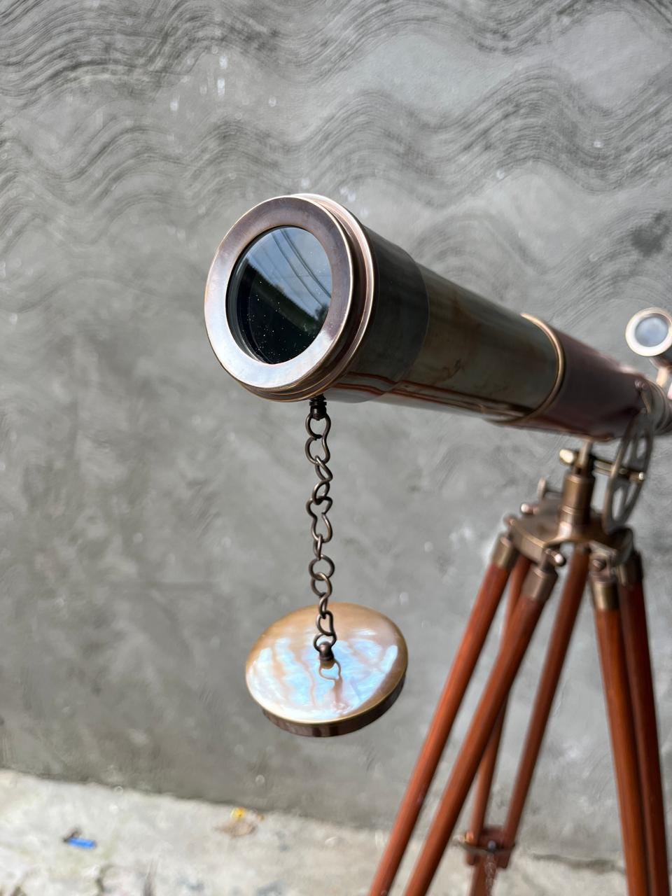 May include: A vintage brass telescope on a wooden tripod. The telescope features a dark brown barrel with brass detailing and a lens cover secured by a chain. The tripod legs are a warm brown colour.