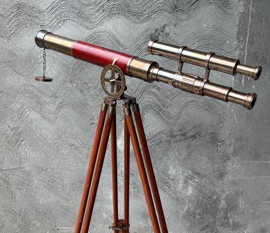 May include: A vintage brass telescope with a red barrel and a secondary scope, mounted on a wooden tripod. The telescope features a small chain with a weight and a brass adjustment wheel. The tripod legs are made of dark wood.