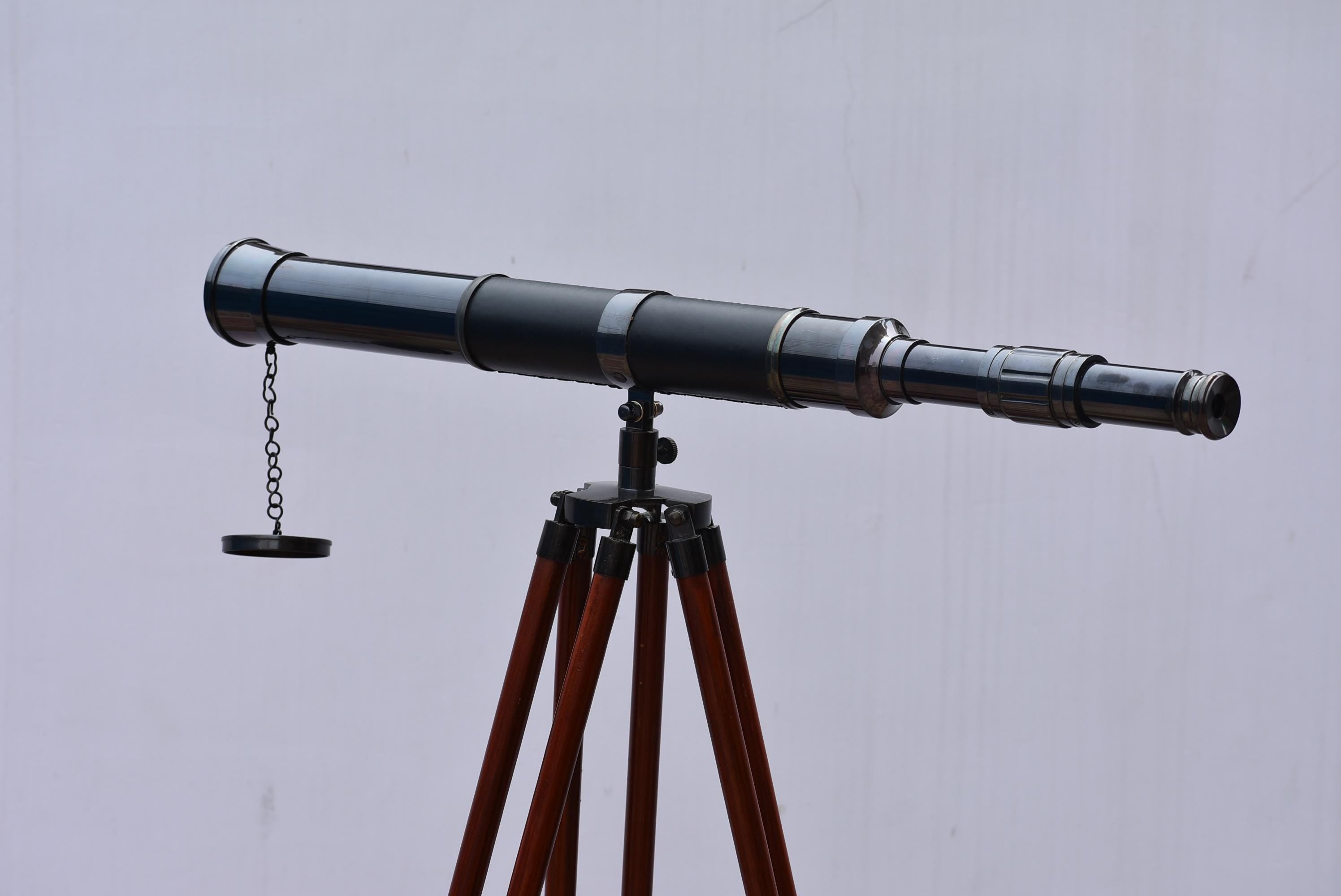 May include: A black telescope on a dark brown wooden tripod. The telescope features a chain and a small cap. The tripod has three legs. The telescope is made of metal and has a leather-like grip.