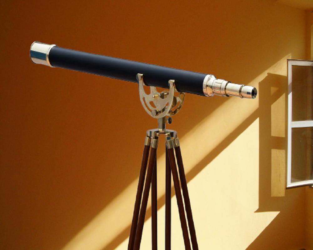 May include: A black telescope with silver accents and a brass-colored mount is mounted on a wooden tripod. The telescope is positioned against a yellow wall, with sunlight casting shadows.
