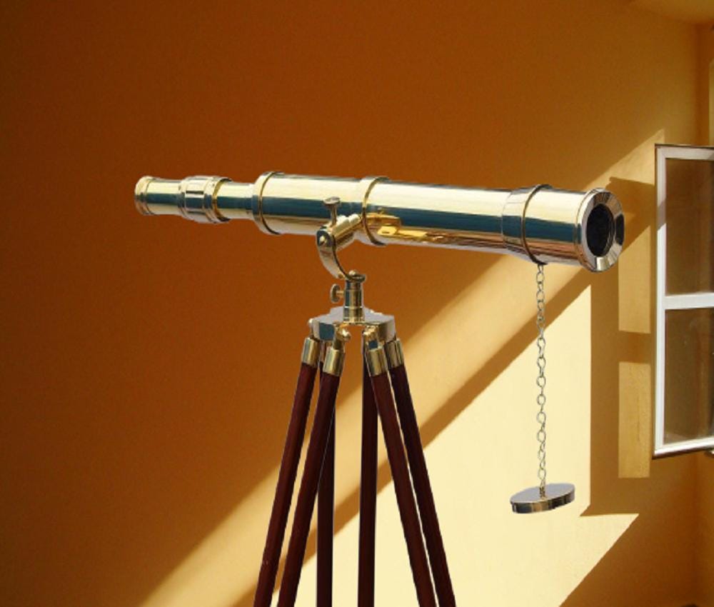 May include: A brass telescope on a dark brown wooden tripod. The telescope has a shiny, metallic finish and is mounted on a three-legged stand. A small chain with a circular weight hangs from the telescope. The background is a warm yellow.