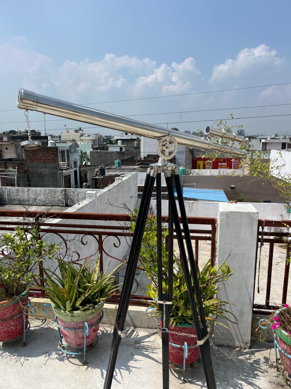 May include: A silver telescope on a black tripod stand, positioned outdoors. The telescope features a long, cylindrical barrel and is mounted on a robust, three-legged stand. The background reveals a cityscape under a blue sky.
