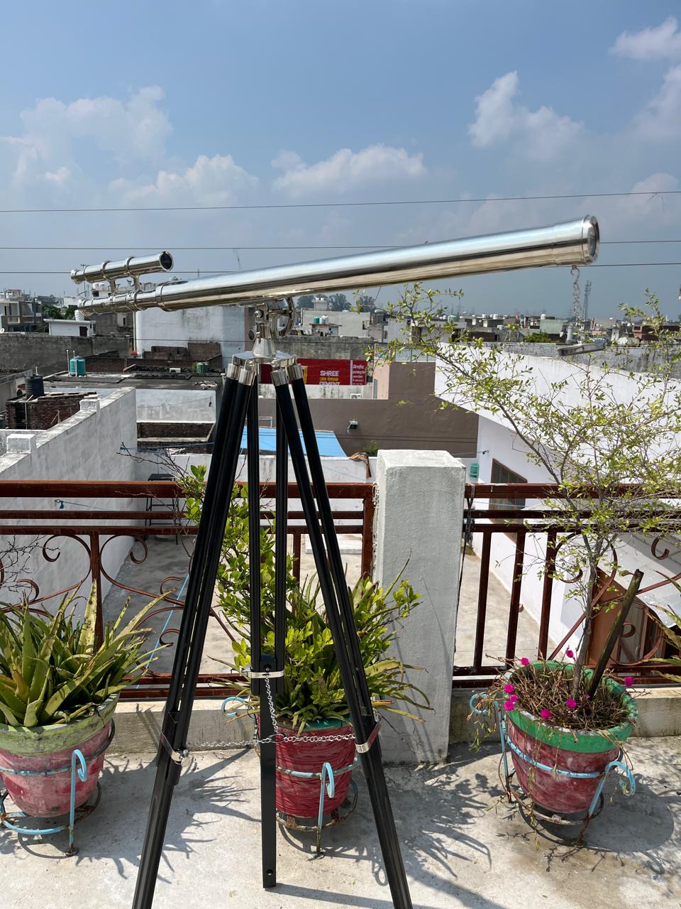 May include: A silver telescope on a black tripod, with a sunny sky and rooftops in the background. The telescope features a long, cylindrical barrel and smaller sighting scopes. Potted plants are at the base.