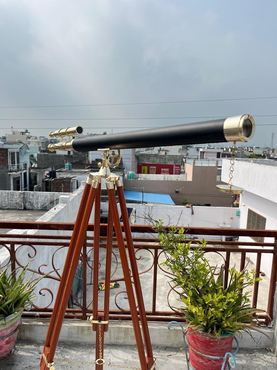 May include: A black telescope with gold-coloured fittings is mounted on a wooden tripod. The telescope has a long barrel and a smaller, angled eyepiece. The tripod has three legs and is made of dark wood. The scene is set outdoors with a cloudy sky in the background.