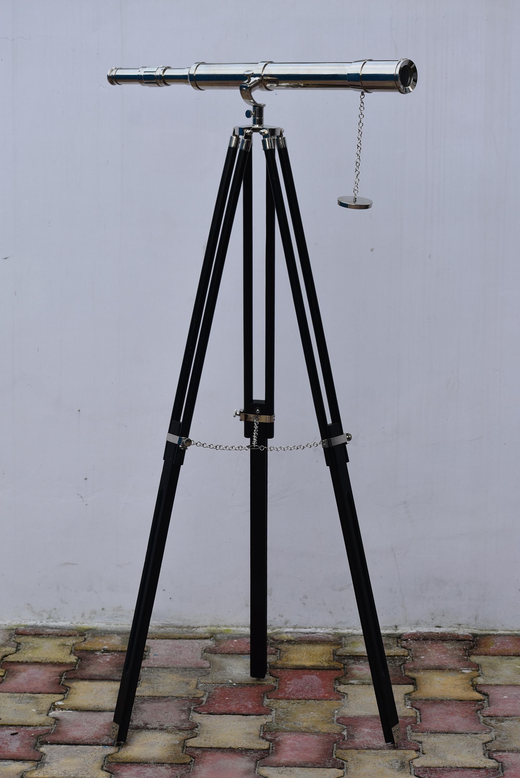 May include: A vintage telescope with a polished metal finish, mounted on a black wooden tripod. The tripod legs are angled and connected with metal hardware and a chain. The telescope is pointed horizontally.