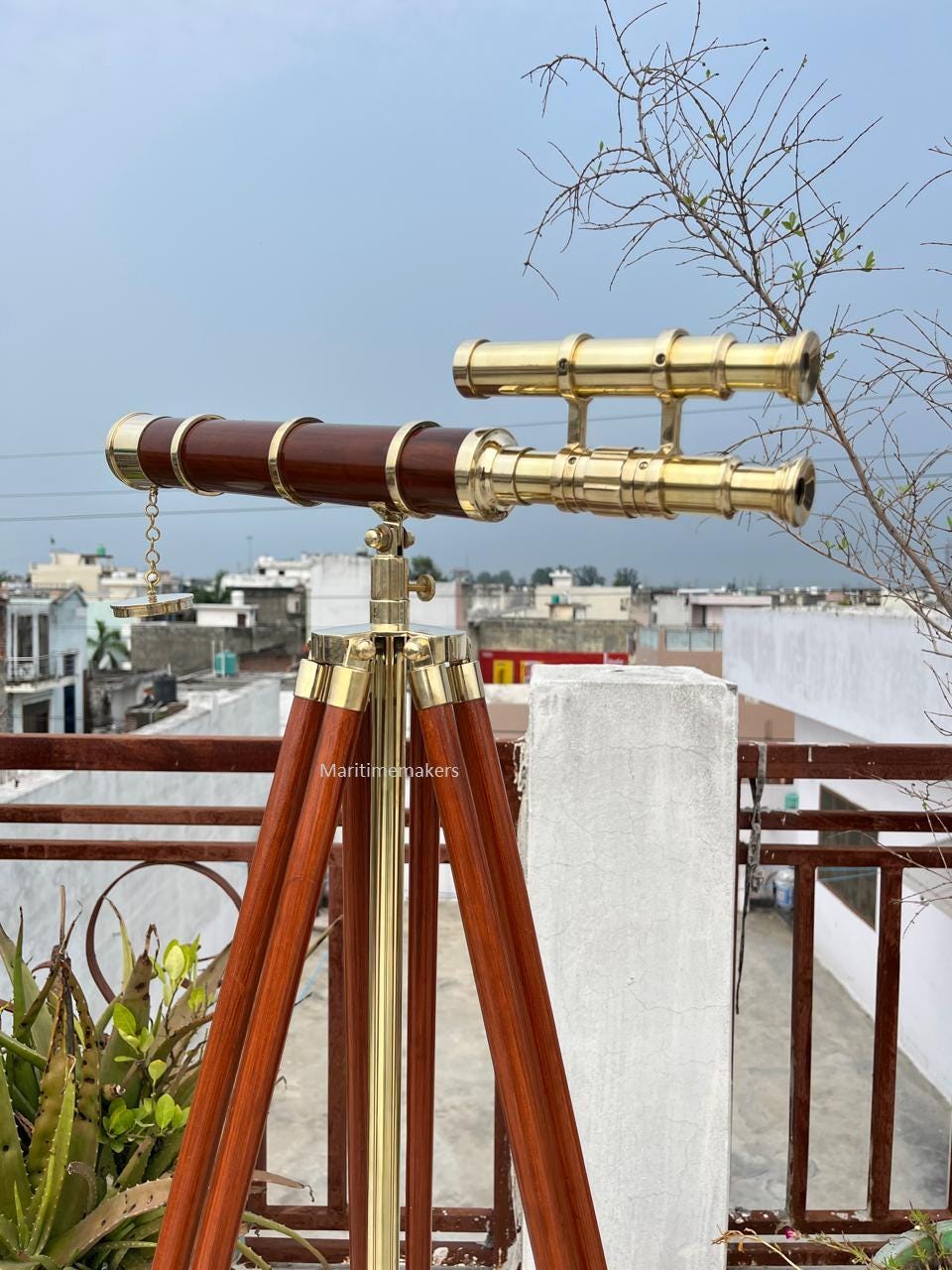 May include: A brass and wood nautical telescope on a wooden tripod. The telescope has a dark brown wooden barrel with brass accents and a chain. The tripod legs are also made of wood. The text "Maritimemakers" is visible on the tripod.