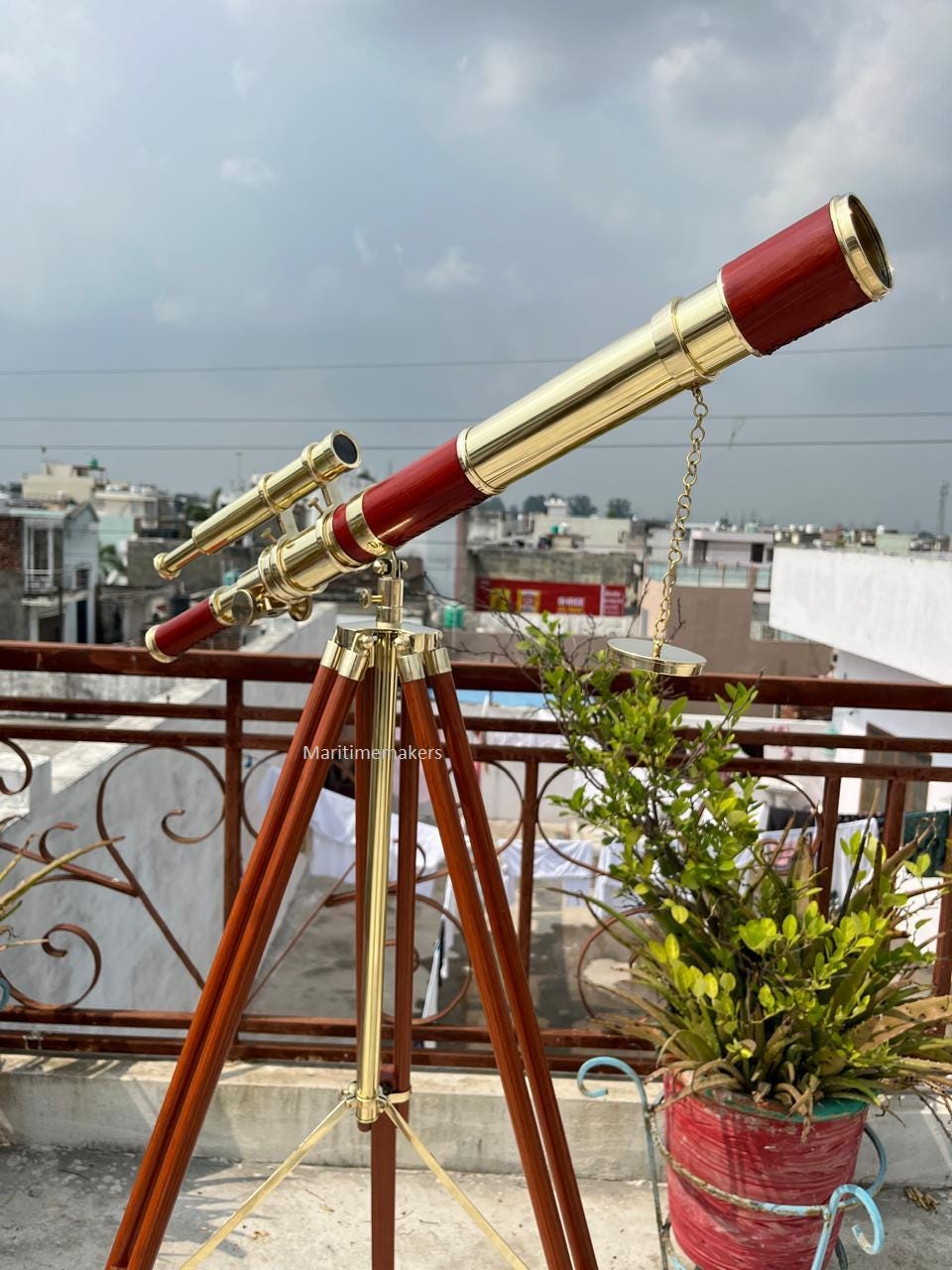 May include: A vintage-style telescope with a brass and red finish, mounted on a wooden tripod. The telescope has a long barrel and a smaller secondary scope. The tripod legs are dark brown, and the metal components are gold-colored.