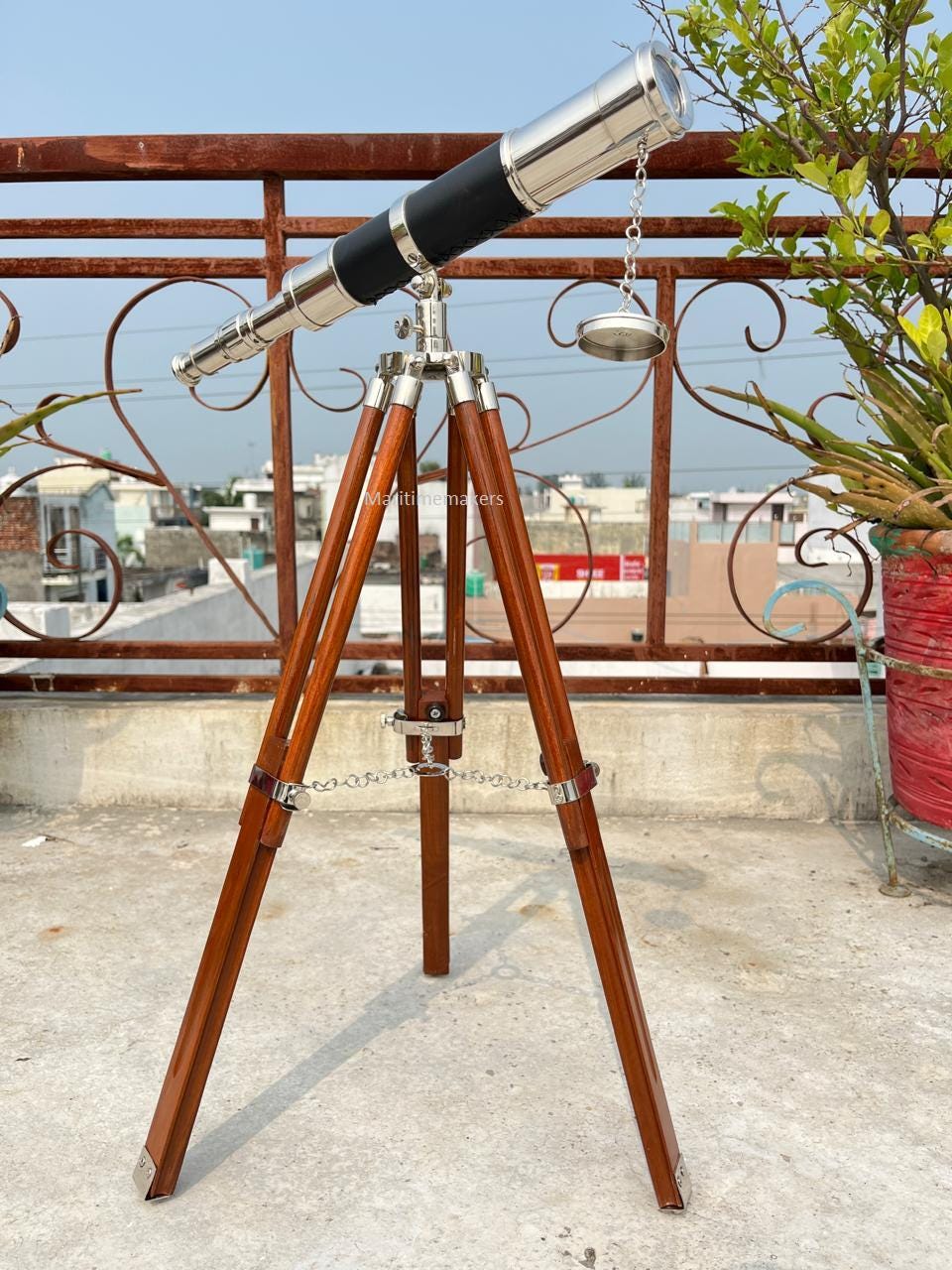 May include: A vintage-style telescope with a shiny silver barrel and black grip is mounted on a wooden tripod. The tripod has dark brown legs with metal accents and a chain. The telescope is aimed upwards, suggesting a focus on celestial observation.