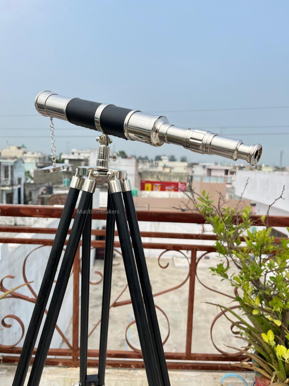 May include: A vintage-style telescope with a silver finish and black accents, mounted on a black tripod. The telescope has a chain and is positioned outdoors. The tripod legs are black and the metal hardware is silver.