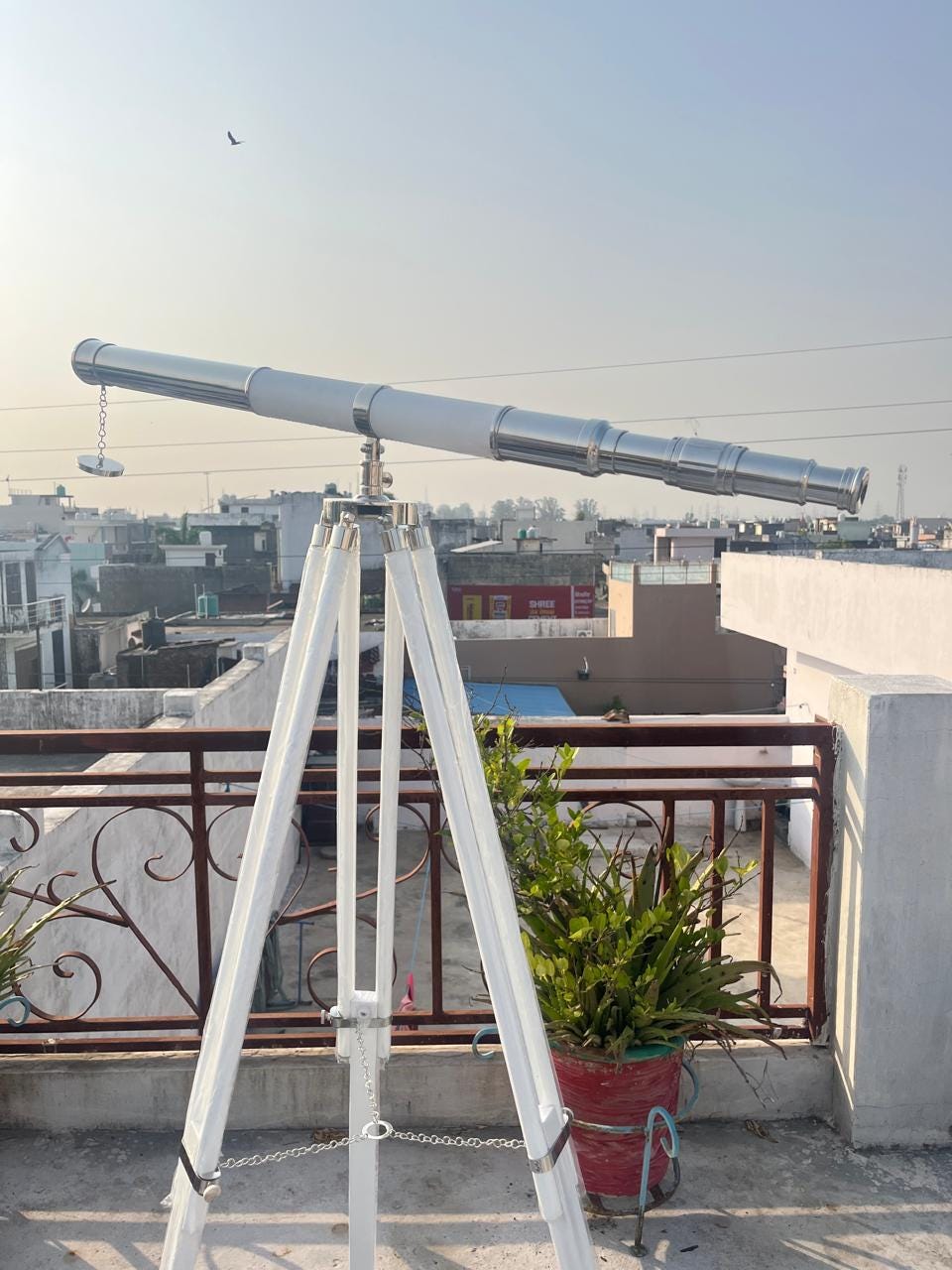 Può includere: Un telescopio argentato e bianco montato su un treppiede di legno bianco. Il telescopio ha una canna lunga con più sezioni e una catena appesa lateralmente. Il treppiede è posizionato su un tetto con un paesaggio urbano sullo sfondo.