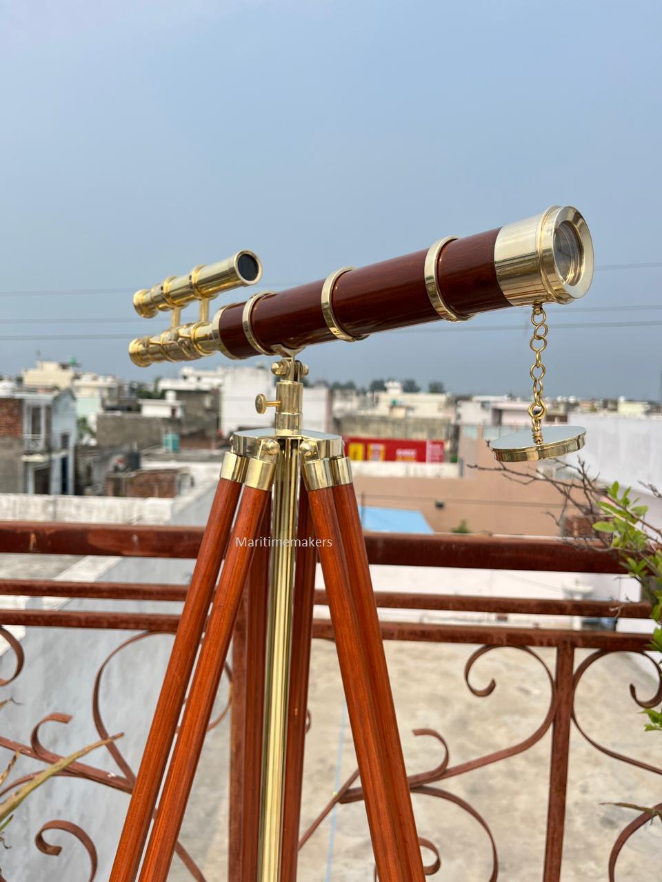 May include: A classic telescope with a dark brown barrel and brass detailing. It is mounted on a wooden tripod with brass fixtures. The image shows the telescope outdoors, with a cityscape visible in the background.