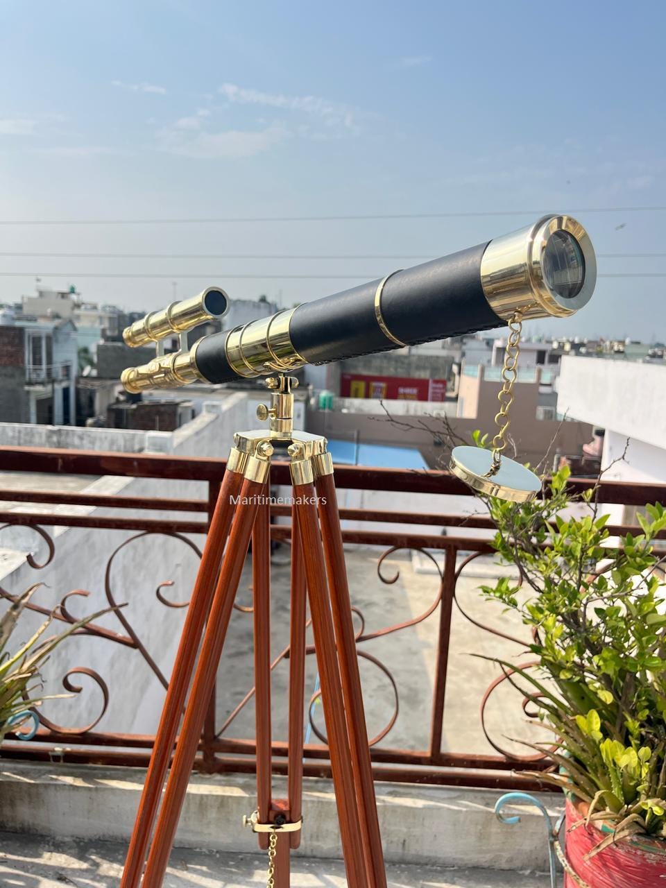 May include: A brass and black telescope on a wooden tripod. The telescope features a black leather-wrapped barrel with brass detailing and a lens. The tripod is made of dark brown wood and has brass fittings. The telescope is positioned outdoors.