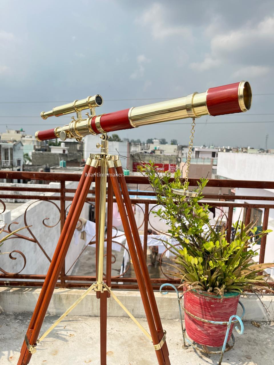 May include: A brass and red telescope mounted on a wooden tripod. The telescope has a long barrel with a red section and a smaller, angled scope on top. The tripod has brass accents.
