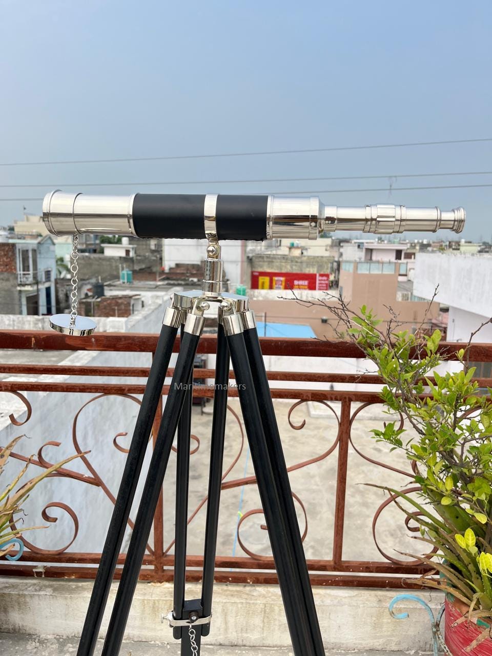 May include: A silver and black nautical telescope mounted on a black wooden tripod. The telescope has a black leather grip and a chain. The tripod has adjustable legs. The background shows a cityscape and a blue sky.