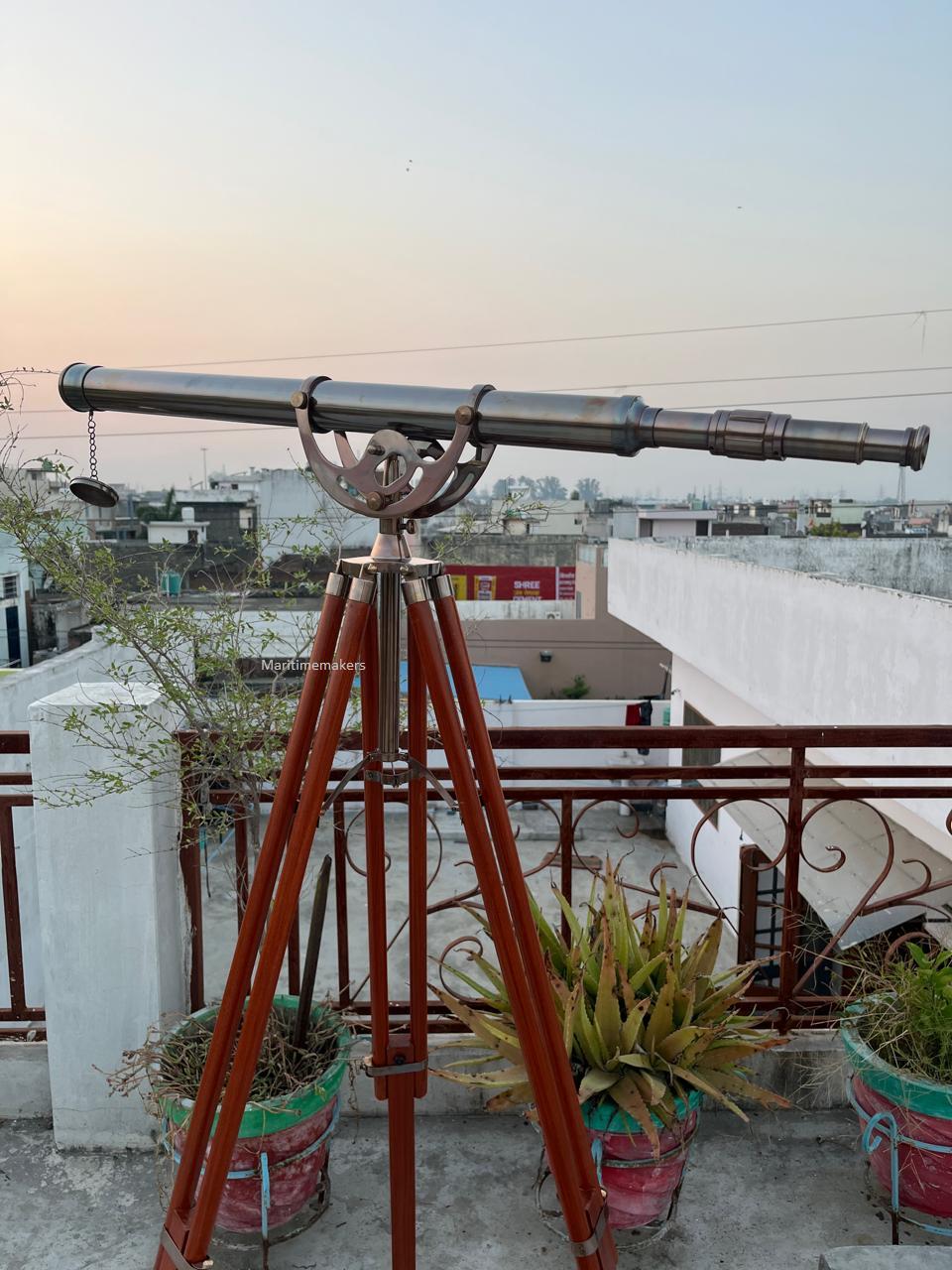 Può includere: Un telescopio in stile vintage montato su un treppiede in legno. Il telescopio è in metallo argentato e ha una lunga canna. Il treppiede ha tre gambe in legno marrone. La scena è ambientata all'aperto con un paesaggio urbano sullo sfondo.