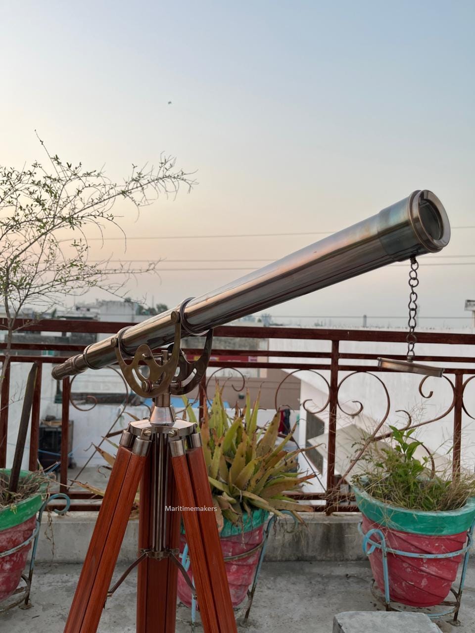 Può includere: Un telescopio in ottone in stile vintage montato su un treppiede in legno. Il telescopio ha una canna lunga e cilindrica con una catena e una lente all'estremità. Il treppiede ha tre gambe in legno. Lo sfondo mostra un tetto con piante in vaso.