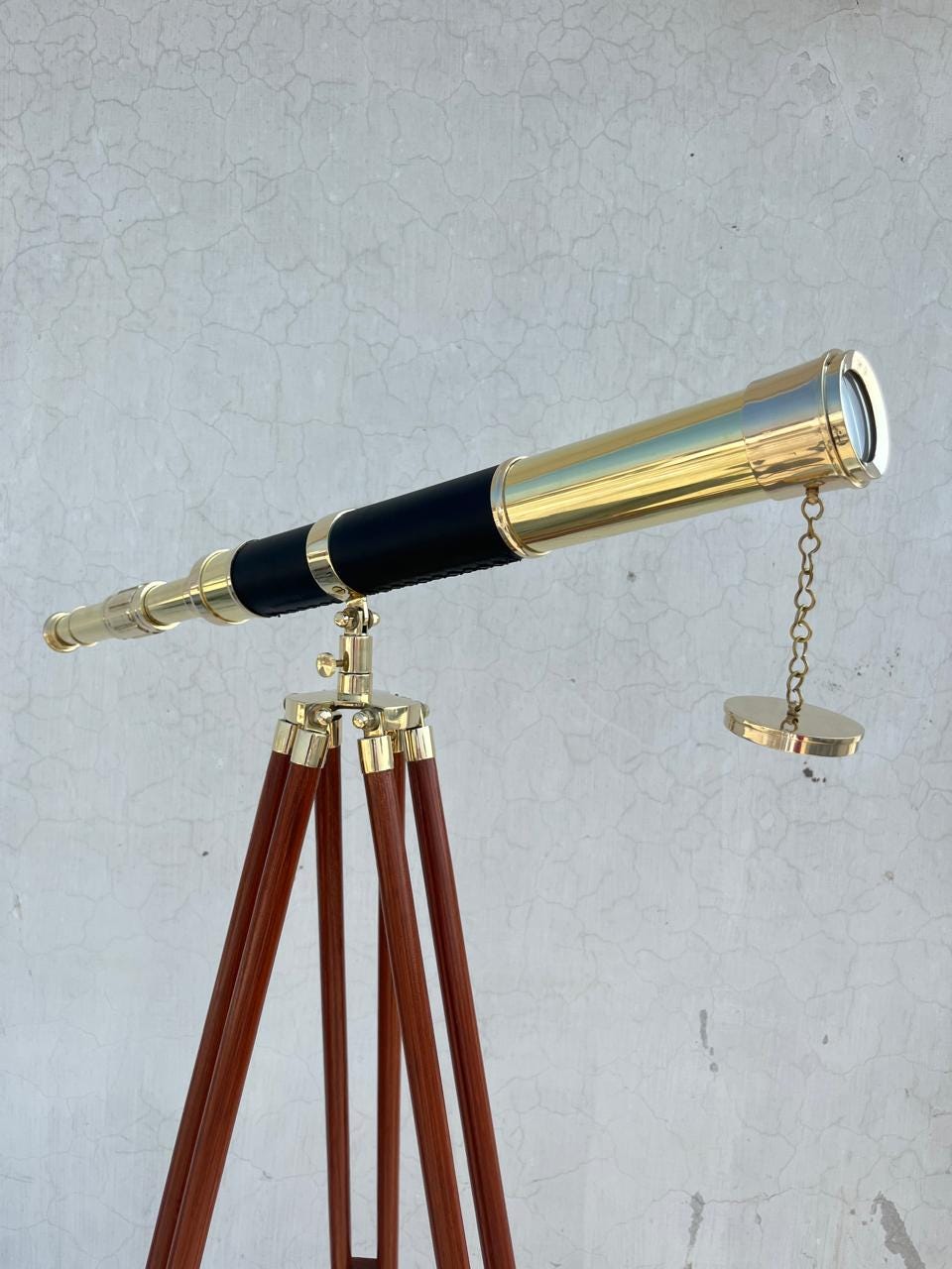 May include: A brass telescope with a black leather grip, mounted on a wooden tripod. The telescope has a gold finish and a lens at the end. The tripod legs are dark brown with brass accents.