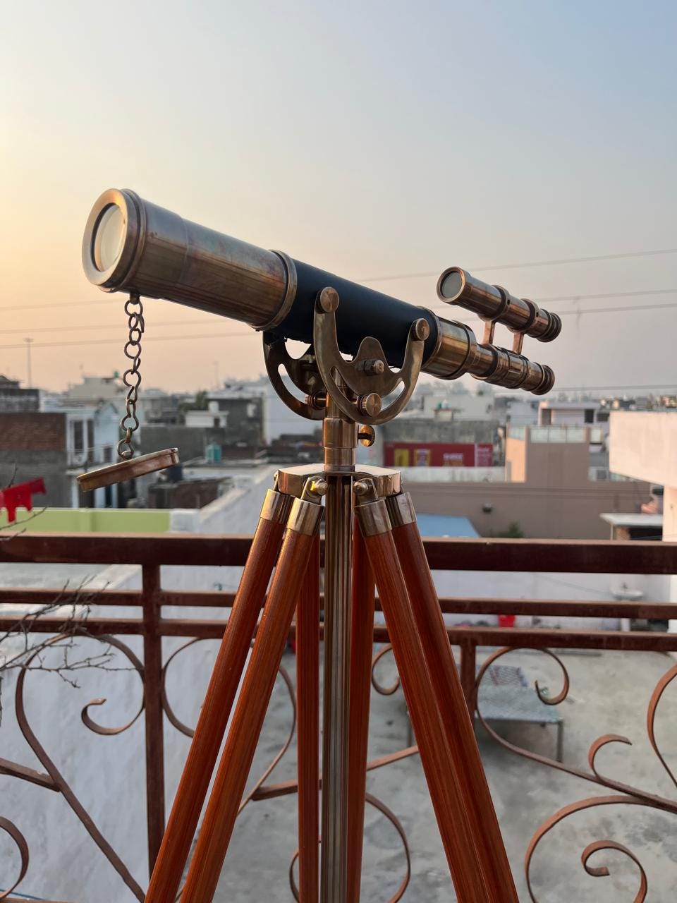 Può includere: Un telescopio in ottone e nero montato su un treppiede in legno. Il telescopio ha una canna lunga con una lente a un'estremità e un mirino più piccolo attaccato. Le gambe del treppiede sono in legno e collegate da supporti metallici.