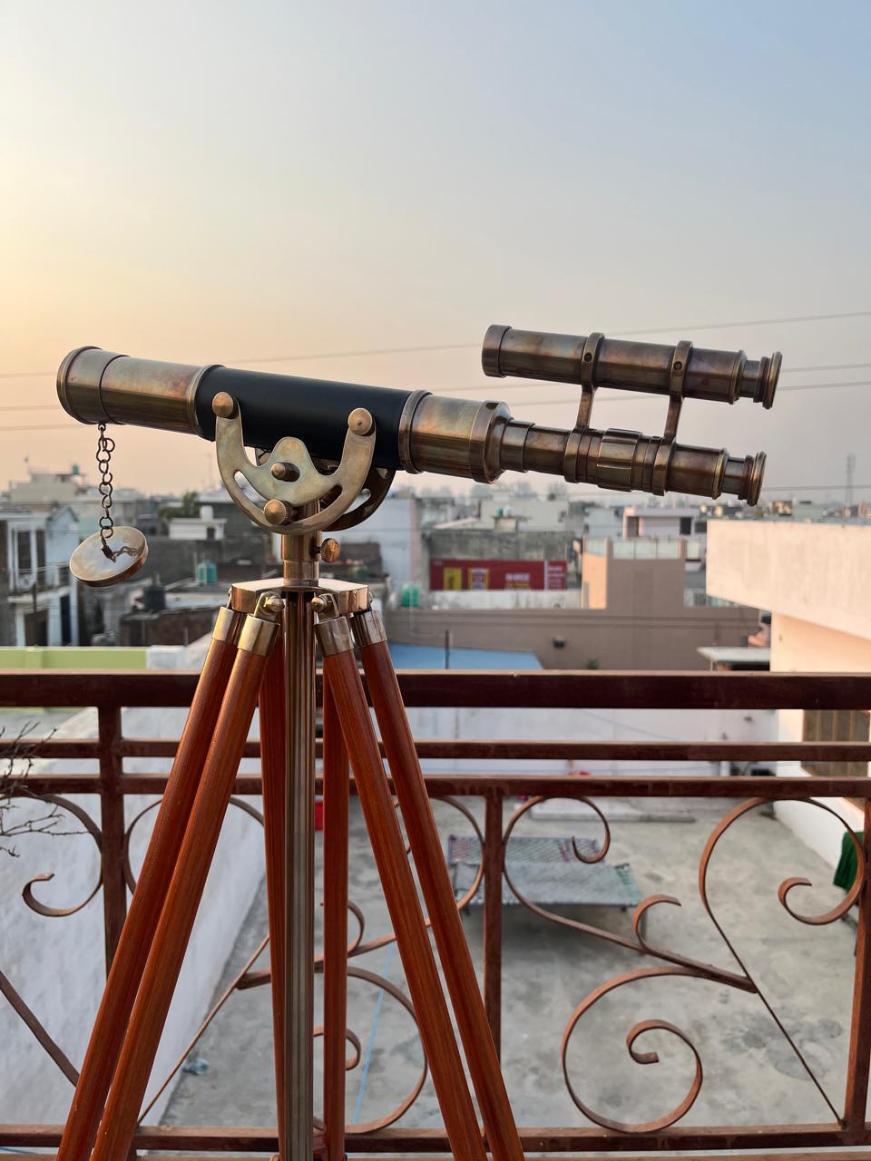 Può includere: Un telescopio in ottone in stile vintage con un tubo centrale nero e un cannocchiale più piccolo e parallelo. È montato su un treppiede in legno con dettagli in ottone. Il telescopio ha una catena e un pezzo circolare.