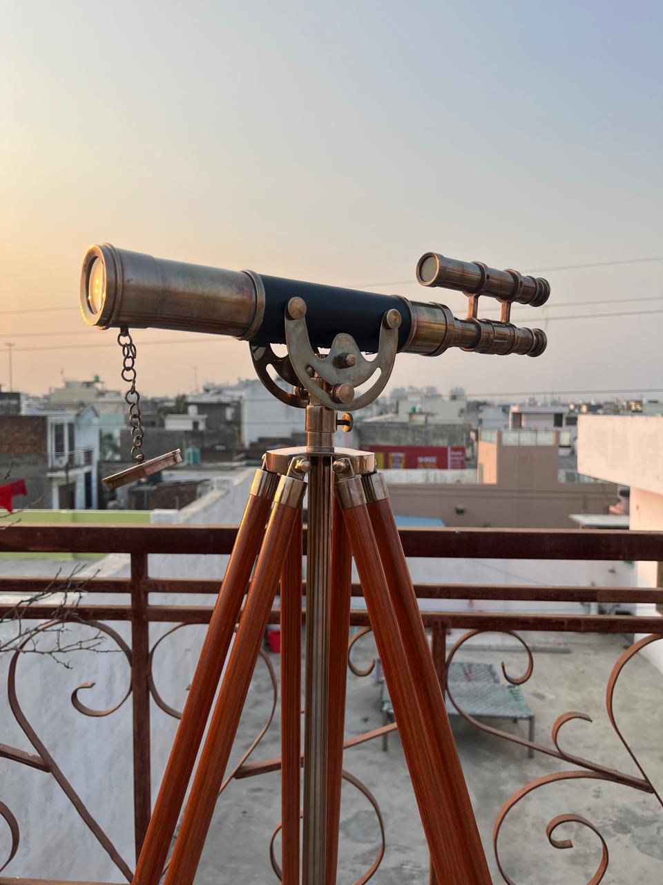 Può includere: Un telescopio in ottone in stile vintage con un corpo nero, montato su un treppiede in legno. Il telescopio ha un mirino secondario e una catena. Le gambe del treppiede sono di colore marrone caldo. Lo sfondo mostra un paesaggio urbano al tramonto.