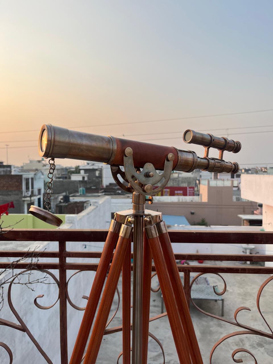 May include: A classic brass telescope with a leather-clad body, mounted on a wooden tripod. The telescope includes a secondary scope and is set up outside, with buildings and a sunset in the background.