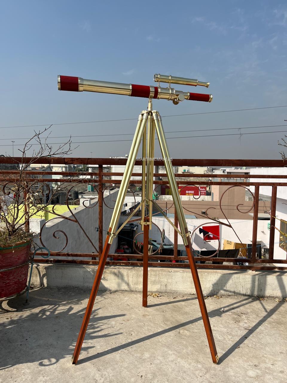May include: A brass and wood telescope with a red accent on a tripod stand. The telescope has a long barrel and a smaller secondary scope. The tripod legs are brown wood with brass hardware. The telescope is set up outdoors.