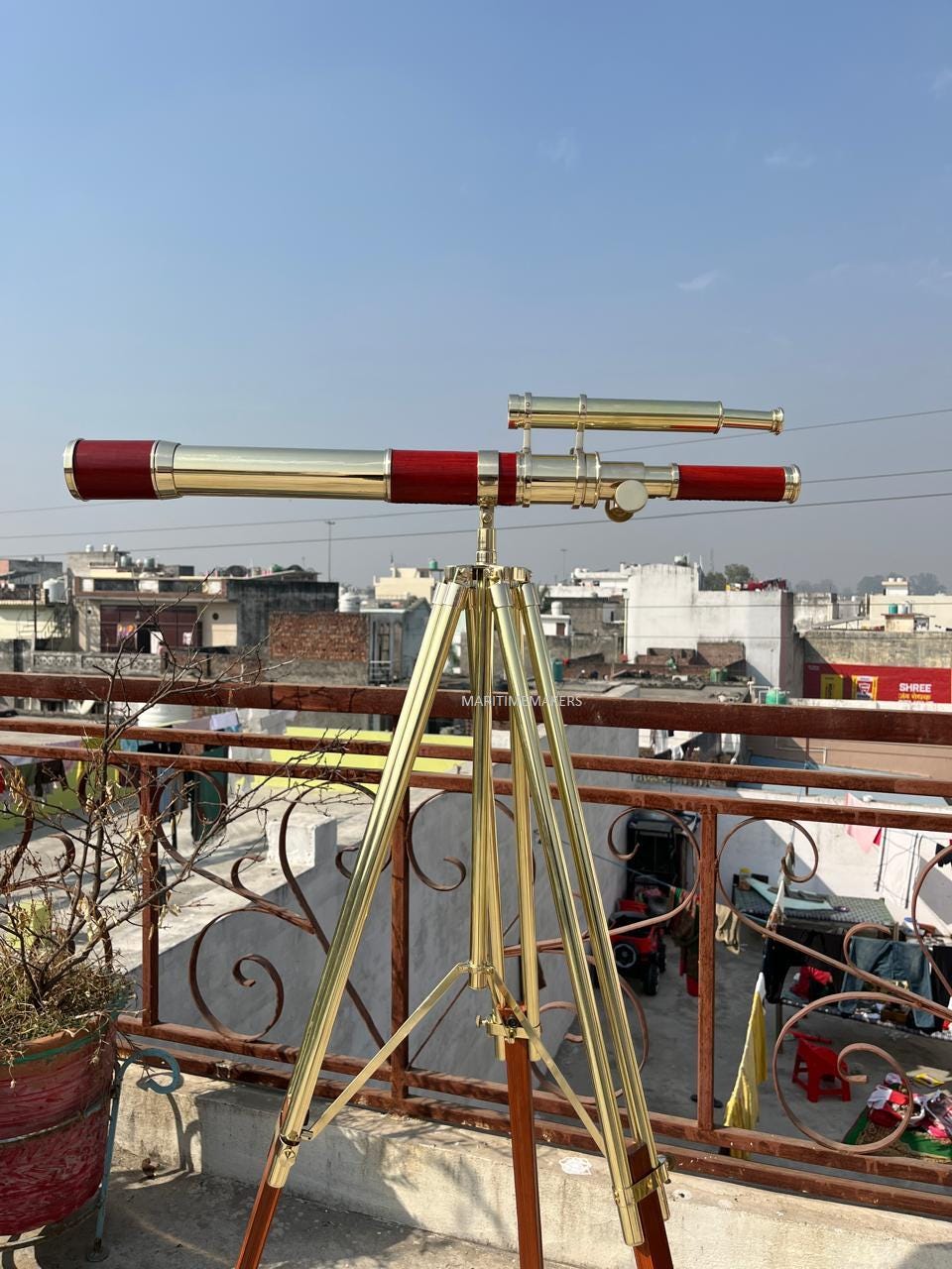 May include: A brass and wood telescope with a red accent, mounted on a brass tripod. The telescope has a long barrel and a smaller, attached scope. The tripod has wooden legs and brass hardware. The image is taken outdoors.