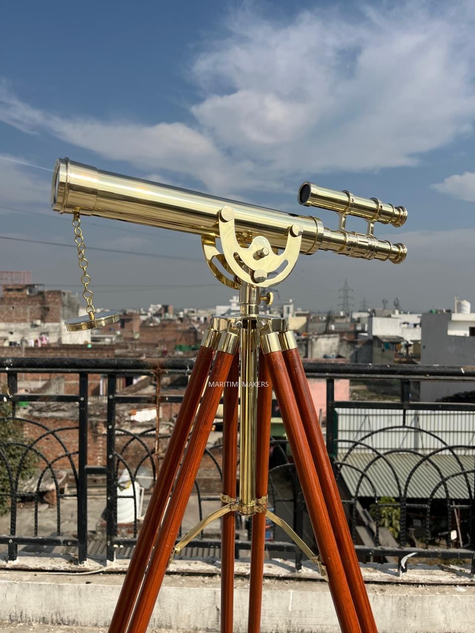 May include: A brass telescope mounted on a wooden tripod. The telescope has a long barrel with a smaller, attached sighting scope. The tripod legs are dark brown, and the metal components are gold-colored. The sky is blue with clouds.