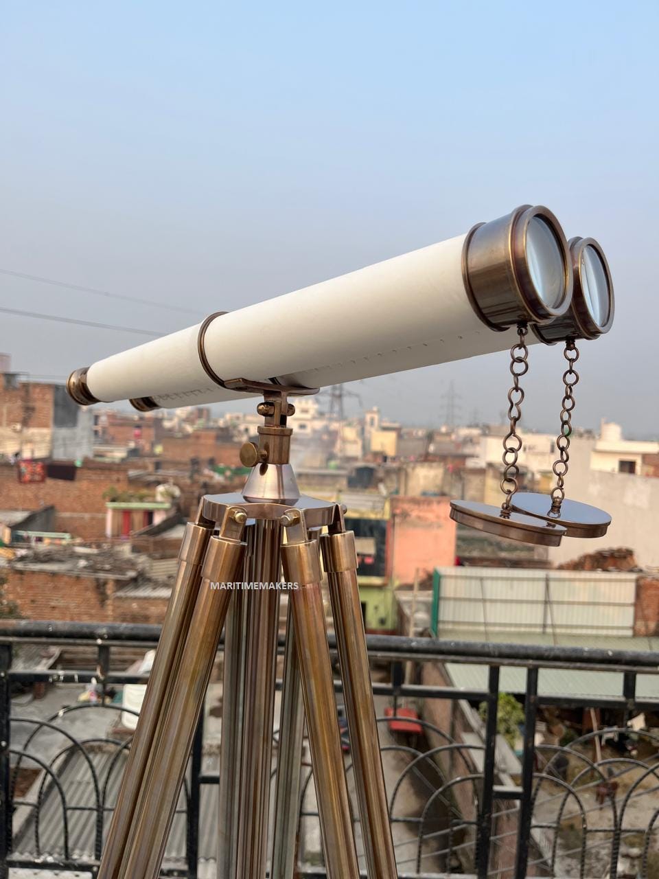 May include: A white and brass nautical telescope on a brass tripod. The telescope features two lenses and is connected to brass covers by chains. The tripod has three legs and is labelled "MARITIMEMAKERS". The background shows a cityscape.