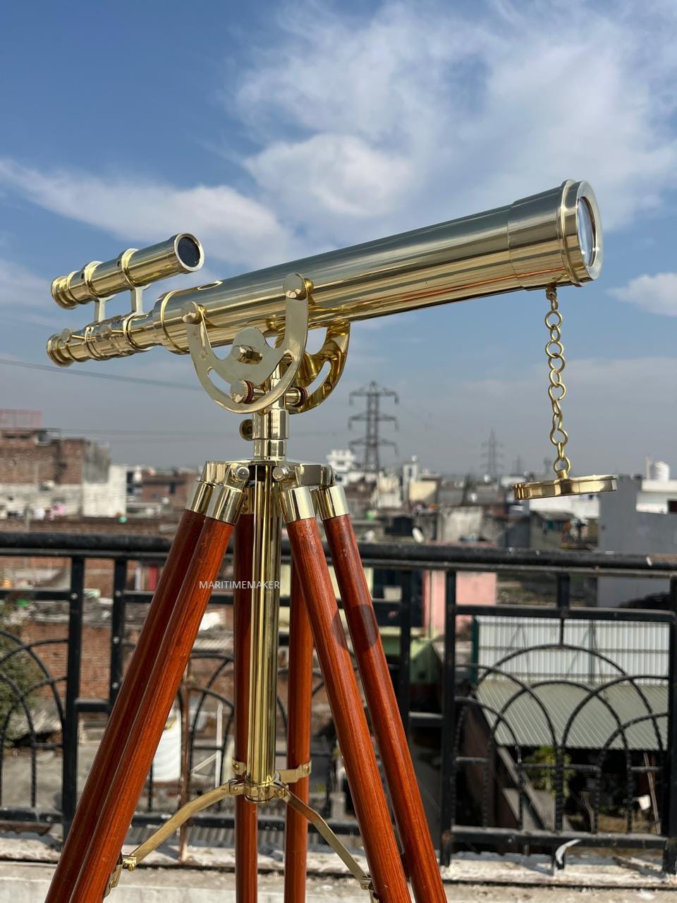 May include: A brass telescope with a wooden tripod. The telescope has a long, cylindrical barrel and a smaller sighting scope. The tripod legs are made of dark wood and are connected by brass hardware. The sky is blue with some clouds.