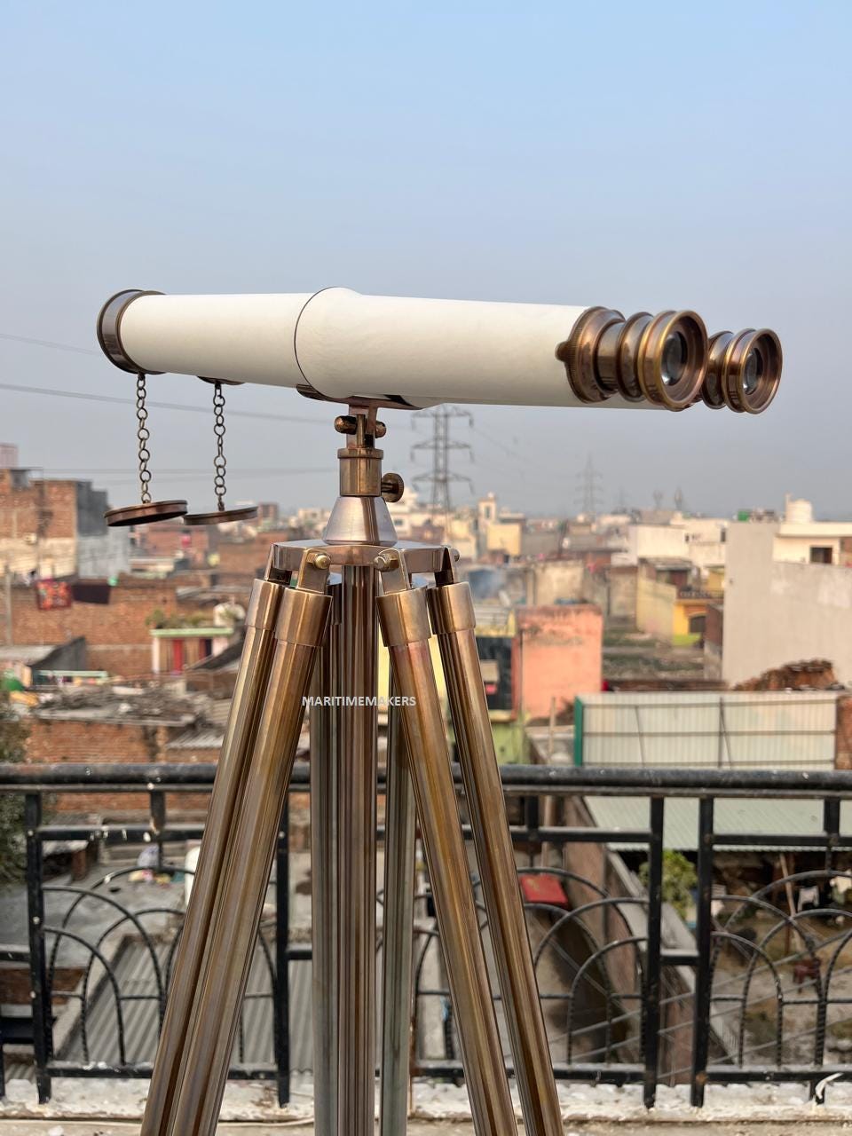 May include: A brass telescope with a white barrel, mounted on a tripod. The telescope has multiple lenses and is connected to the tripod with a brass fitting. The tripod legs are also brass. The background shows a cityscape.